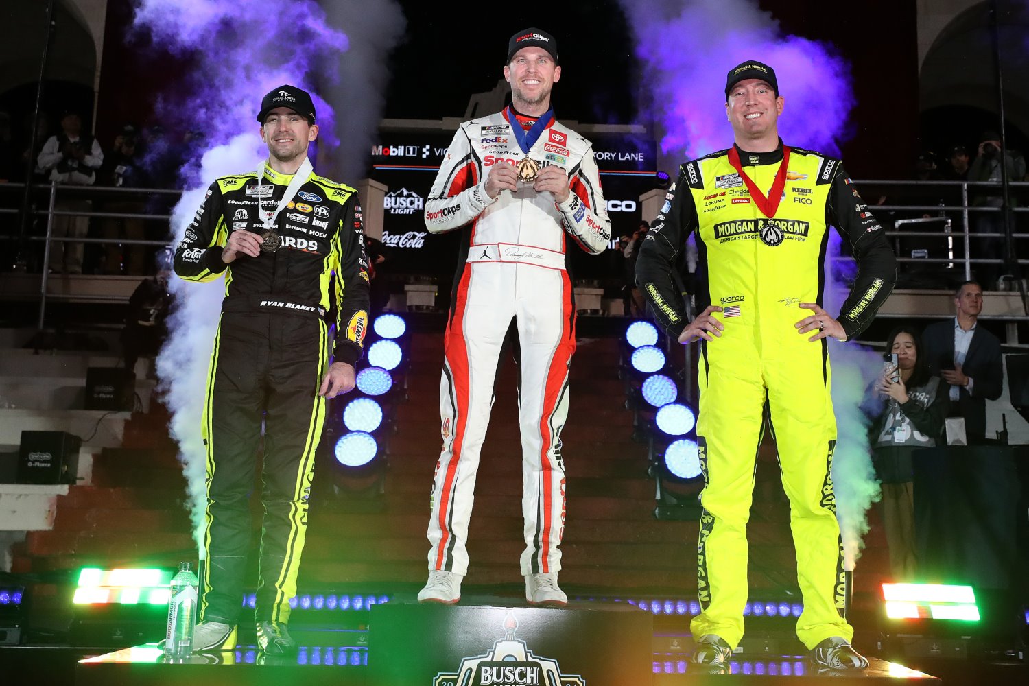 Denny Hamlin, driver of the #11 Sport Clips Haircuts Toyota, winner (C) Kyle Busch, driver of the #8 Morgan & Morgan Chevrolet, (R) second place and Ryan Blaney, driver of the #12 Menards/Great Lakes Flooring Ford, third place pose on the podium in victory lane after the NASCAR Cup Series Busch Light Clash at The Coliseum at Los Angeles Memorial Coliseum on February 03, 2024 in Los Angeles, California. (Photo by Meg Oliphant/Getty Images)